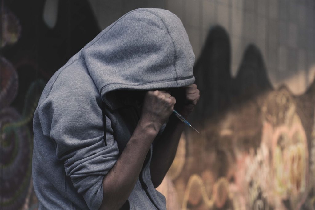 A person in a gray hoodie is crouched with their arms crossed tightly against their face, holding a syringe in one hand. The background is a graffitied wall, casting shadows in a dimly lit environment.