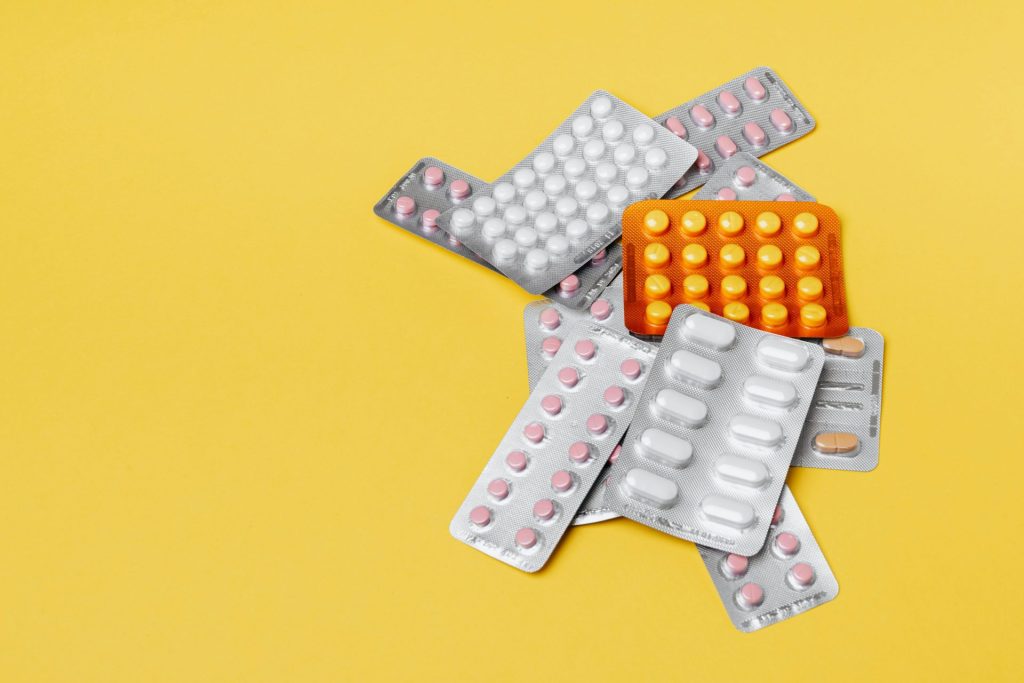 Blister packs of various pills, including pink, white, and yellow tablets, are scattered against a bright yellow background.