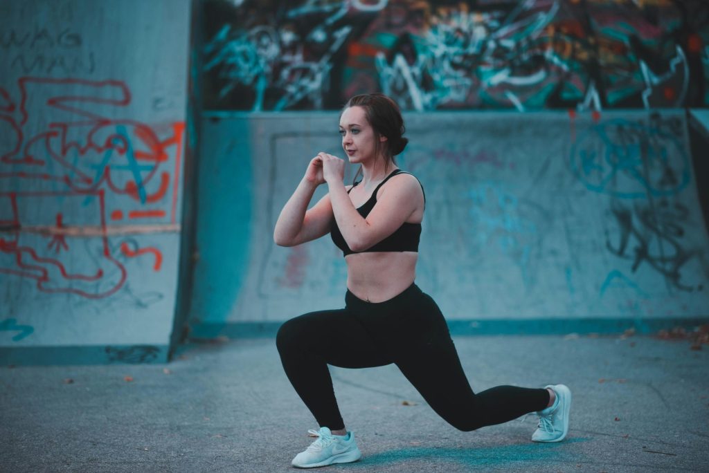 A woman in athletic wear performs a forward lunge on a concrete surface. She is outdoors in front of a graffiti-covered wall, wearing a black sports bra, black leggings, and white sneakers.