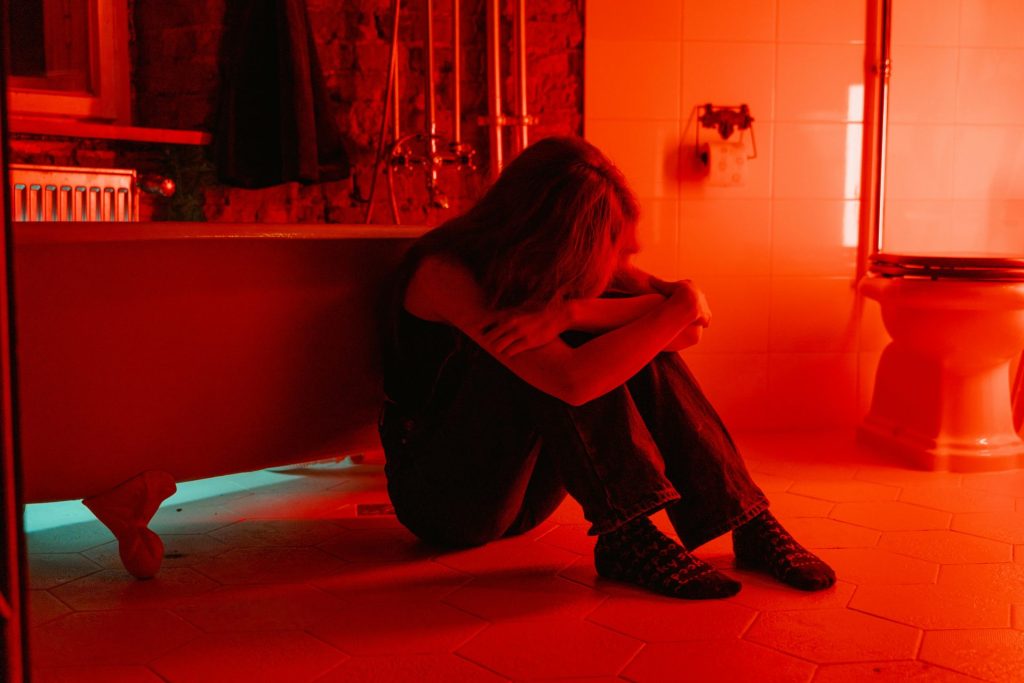 A person sits on the floor of a dimly lit bathroom, leaning against a bathtub with their arms wrapped around their knees. The room is bathed in red and blue lighting, creating a somber atmosphere.