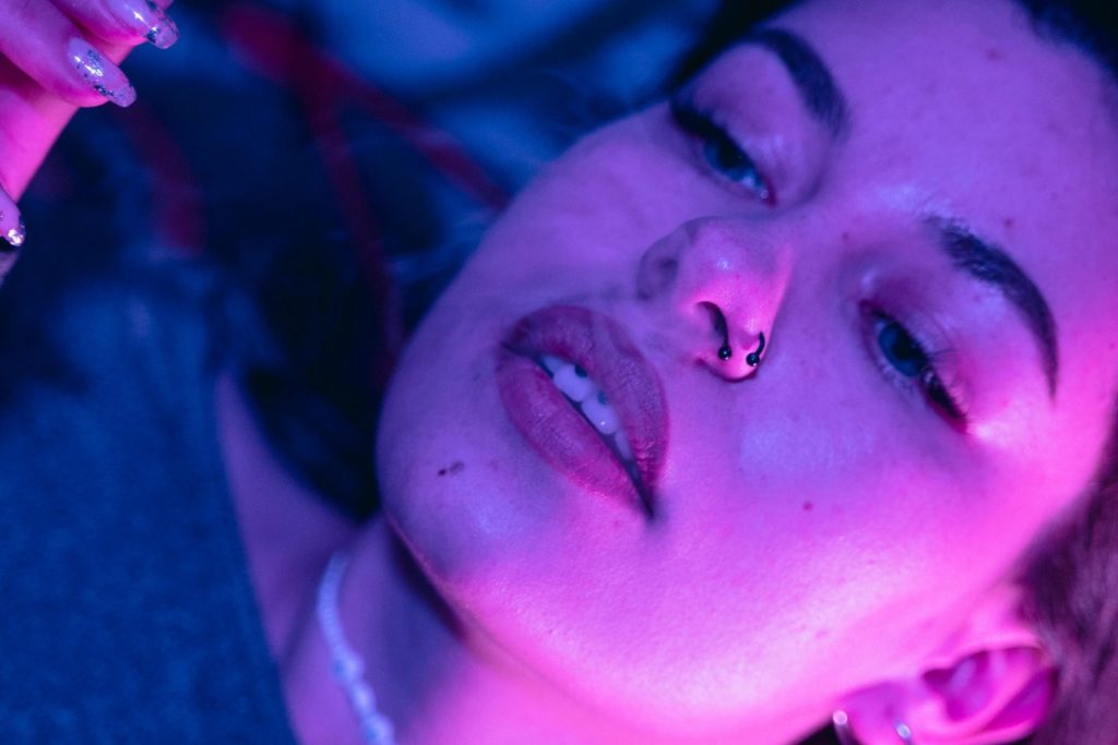 Close-up of a woman under purple lighting, with a nose ring and subtle makeup. Her eyes are looking away from the camera, and she's wearing a necklace. The lighting casts soft shadows on her face.