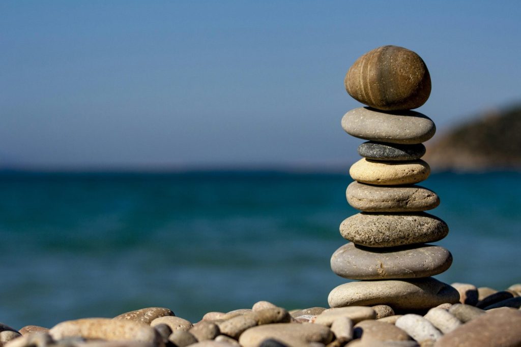 A stack of smooth, round stones balances on a pebble-covered beach. The ocean and a clear blue sky fill the background, creating a serene and tranquil scene.