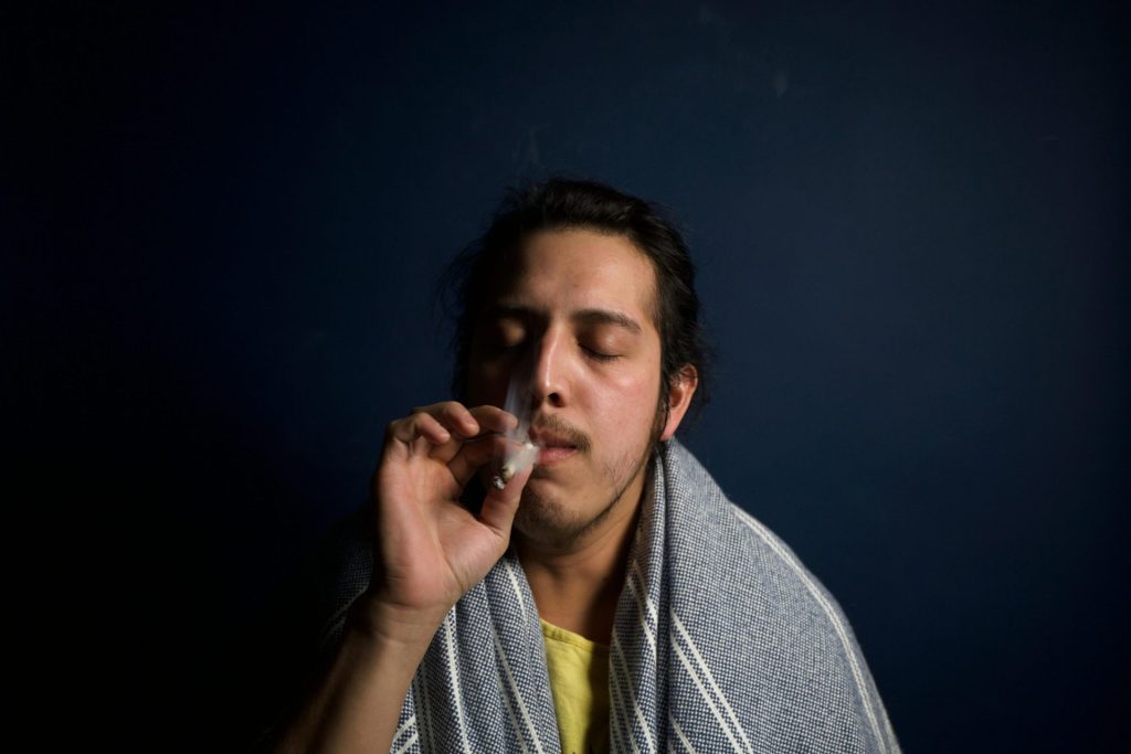 A person with long hair, wrapped in a gray blanket, holds a cigarette to their lips. Their eyes are closed, suggesting relaxation. The background is a dark, plain backdrop, highlighting the subject.