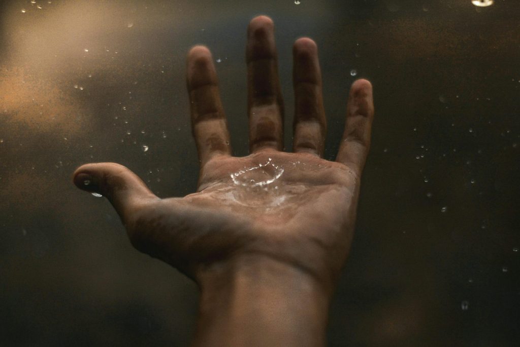 A close-up of an open hand catching a drop of water, creating a splash in the palm. The background is dark and blurred, enhancing the focus on the moment of contact.