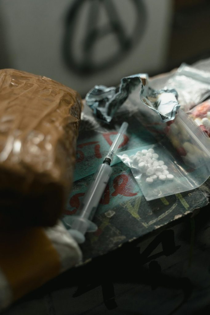 A dimly lit table with a syringe, a package wrapped in brown paper, a small bag of white pills, and crumpled foil. A partially visible anarchy symbol in the background.
