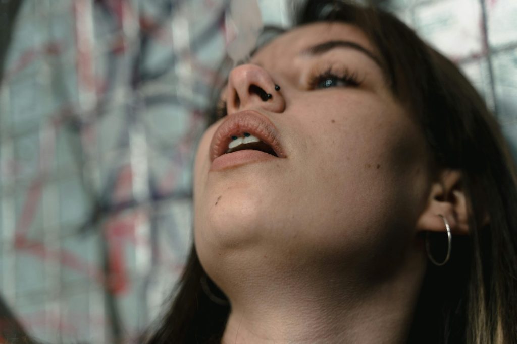 A close-up of a person with long hair, looking upward. They have a nose ring and are wearing hoop earrings. The background features a blurred graffiti wall with various colors.