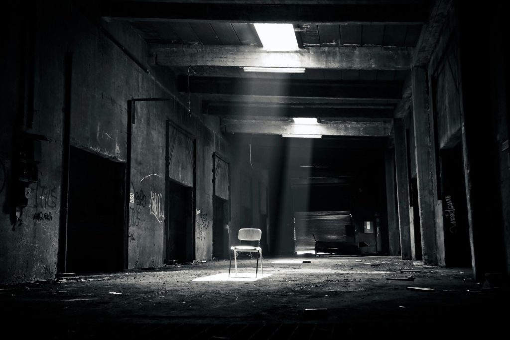 A solitary chair sits in the center of an abandoned, dimly lit industrial warehouse. Sunlight streams through narrow openings in the ceiling, casting light on the dusty floor and highlighting graffiti on the walls.