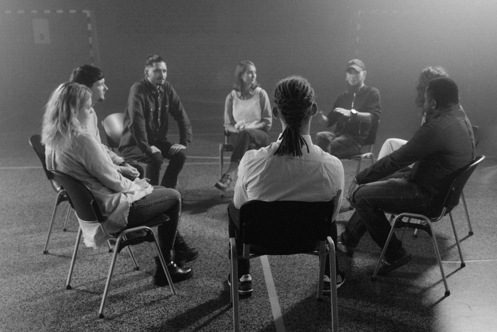 A black and white image shows a group of people sitting in a circle on chairs in a spacious room. They appear to be engaged in a discussion or meeting. The atmosphere is subdued, with soft lighting and a slightly foggy background.