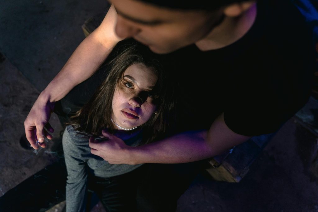 A woman with long hair looks up while sitting on the ground, leaning against a man's lap. She wears a gray top and the scene is dimly lit, suggesting an intimate or contemplative atmosphere. The man is dressed in a black shirt.
