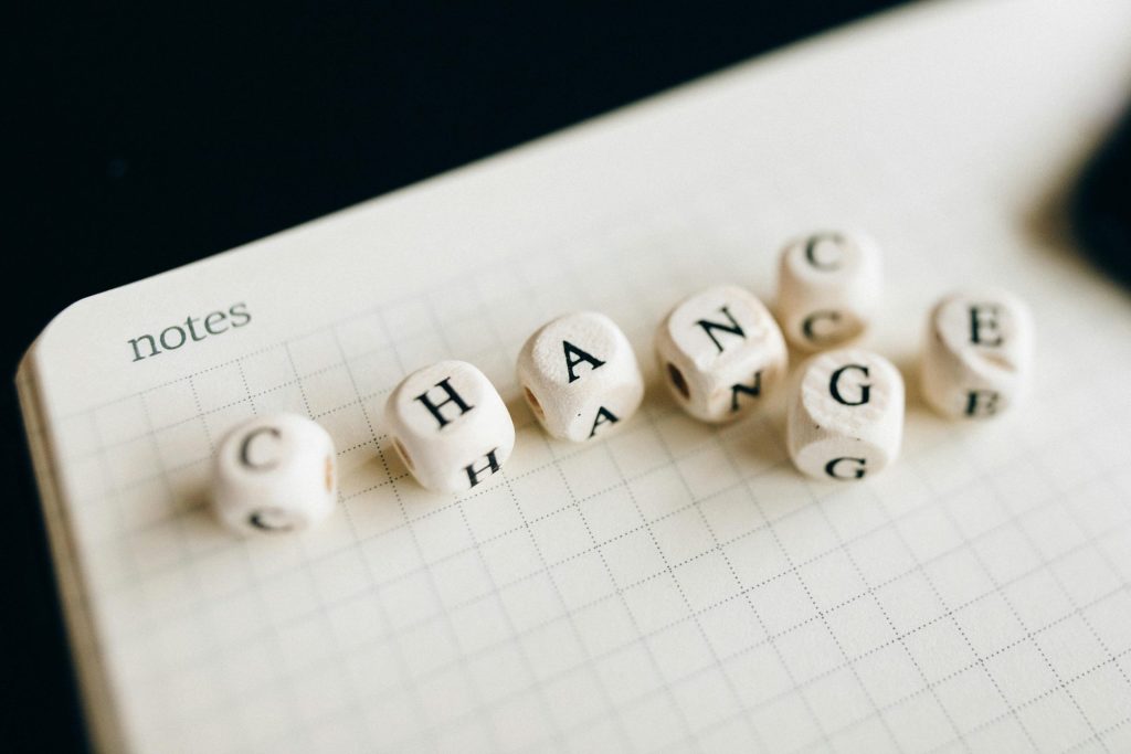 Wooden dice with letters spell "CHANGE" on graph paper labeled "notes." Some dice are slightly misaligned, with "G" and "E" tilted. The setup symbolizes transformation or adaptation.